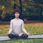 Good-looking girl in sports clothing is twisting her body sitting in lotus position then relaxing with closed eyes after practice in town park on autumn day.