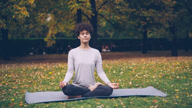 Good-looking girl in sports clothing is twisting her body sitting in lotus position then relaxing with closed eyes after practice in town park on autumn day.